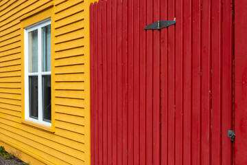 A bright yellow wooden vertical clapboard siding on the exterior wall of the building. There's a white trim closed glass window in the middle. Attached is a deep red wooden gate with black metal hinge