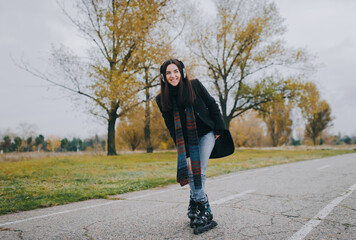 Fototapeta premium Young beautiful smiling girl in casual wear roller skating listens to music. Autumn walk in the fresh air in cloudy weather.