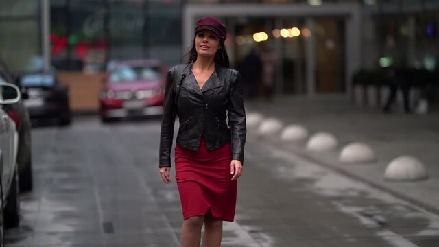 beautiful business confident woman in a purple cap and leather jacket and red skirt strolling on a city street, smiling.