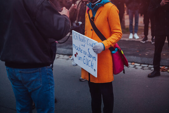 Woman in bright orange coat holding a sign is speaking with a demonstrator on a conspiracy theory demonastration. Her sign says "I had Covid-19 an I am worried about you".