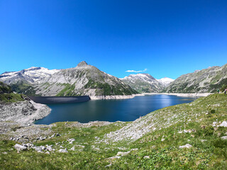 Naklejka na ściany i meble A panoramic view on an artificial, dam lake stretching over a vast territory around Alps in Austria. Lake is shining with navy blue color. In the back there are a few glaciers. Controlling the nature