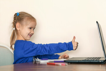 Little girl in a blue blouse raised her finger up. The child sits in front of the laptop and points to the monitor thumbs up