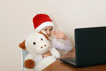 Little girl in a Santa Claus hat with a soft toy. Little girl communicates with friends remotely on the computer. Online Christmas celebration.