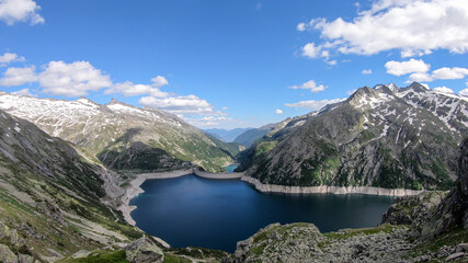 Obraz na płótnie Canvas A panoramic view on an artificial, dam lake stretching over a vast territory around Alps in Austria. Lake is shining with navy blue color. In the back there are a few glaciers. Controlling the nature