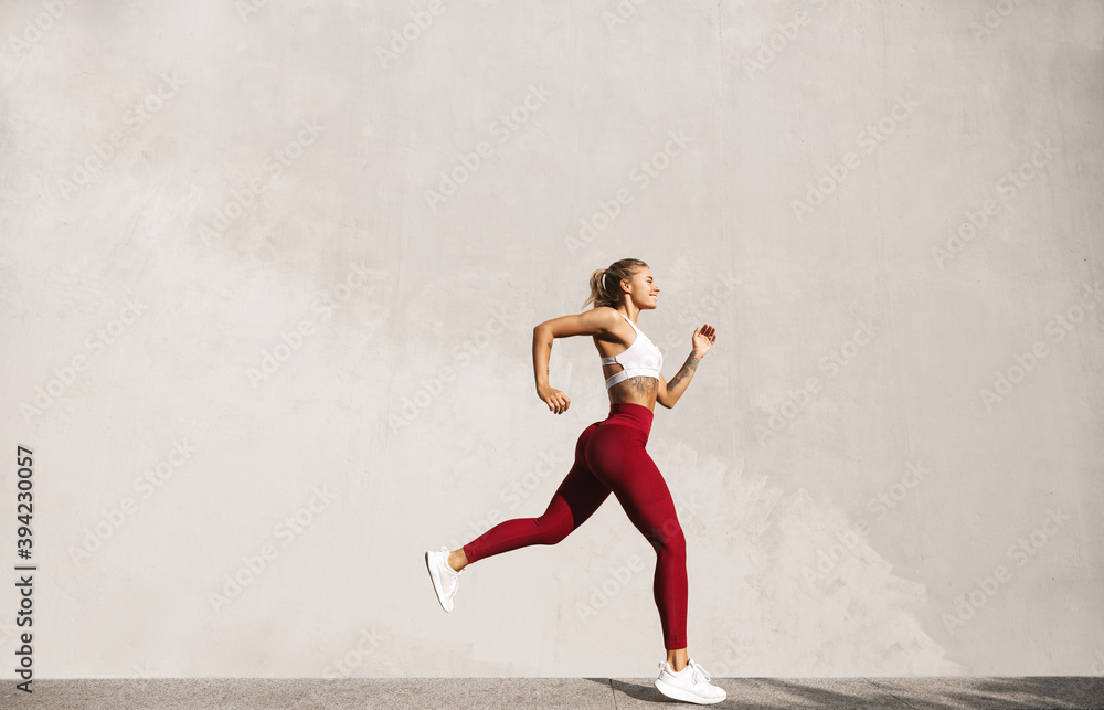 Wall mural Healthy young woman running in morning. Fitness model exercising outdoors. Full length side view shot