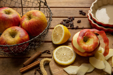 Making of an apple pie: Wooden table with a basket of beautiful red apples, one peeled and some sliced apples, and a cut up lemon. Raisins, cinnamon and Pie dishes in the background.