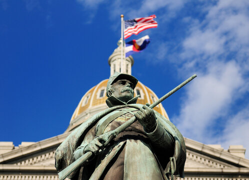 Bottom Up View To The Statue Of A Soldier In Front Of The Shimmerig Golden Dome Of The Colorado State Capitol In Denver On A Sunny Summer Day With A Clear Blue Sky And A Few Clouds