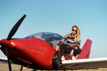 Young woman model with a modern haircut and fashionable sunglasses posing near a red plane wearing trendy casual outfit and black backpack