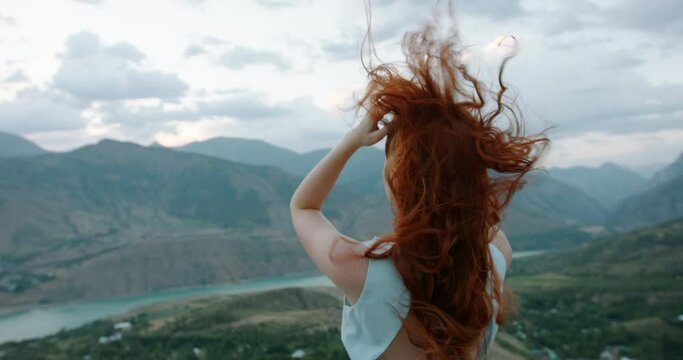 Gorgeous caucasian girl with red hair looking at scenic view from top of a mountain. Wind cinematically blowing hair and white dress - tranquility, peace, freedom 4k footage