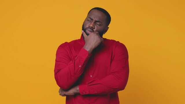 Puzzled pensive young bearded african american man 20s wearing basic red shirt posing isolated on yellow color background in studio. People lifestyle concept. Looking aside up put hand prop up on chin