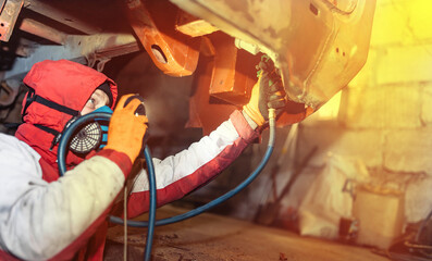 male worker in a respirator treats the underside of the car with anti-corrosion paint with a...