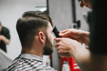 Closeup photo, barber creates a stylish hairstyle for a bearded man, uses a trimmer. Male barber...
