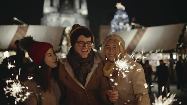Laughing group of friends having fun at the Christmas Fair. People lighting sparklers during winter holidays on the Xmas Market. In the background Santa Claus riding in a sleigh with reindeer.