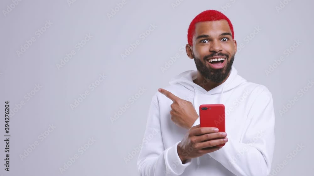 Wall mural Shocked african american man with red hair in streetwear hoodie isolated on white background studio. People lifestyle concept. Using mobile phone typing sms message point finger aside showing thumb up