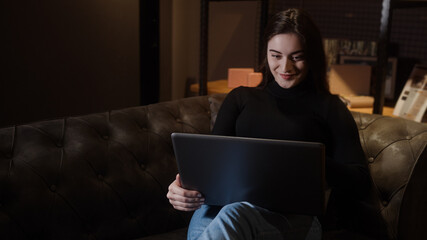 Beautiful charming woman sitting on sofa in her home is using laptop on her lap