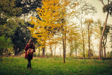 Obraz premium young woman with red coat and leather backpack while walking in the middle of a city park with autumn colors
