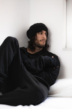 Chico joven guapo vestido de negro con gorro de lana sentado en una cama blanca, mirando hacia un lado, con barba y pelo largo