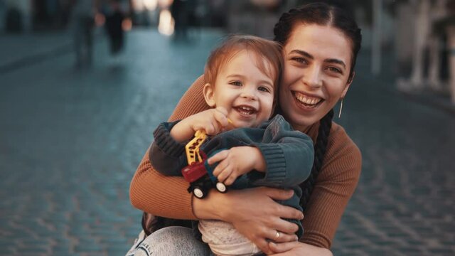 Young happy mother with her cute little child hugs on the city street, look at the camera, smile and laugh, enjoy each other's company, have a great weekend, happy childhood and motherhood, parenting