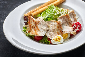 Caesar salad with chicken meat, lettuce, tomatoes, cheese and eggs, on a white plate, on a black background