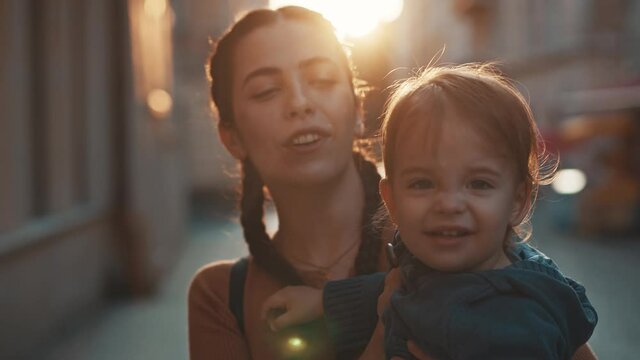 Small very active child in the arms of a young cute mom, kid looks at the camera, smiles and points a finger, mommy straightens baby hair, plays with him and kisses on the cheek, happiness, parent