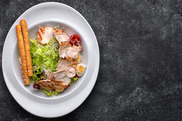 Caesar salad with chicken meat, lettuce, tomatoes, cheese and eggs, on a white plate, on a black background