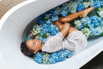 Obraz na płótnie Canvas Woman relaxing in bath with tropical blue hydrangea. Beautiful gently caucasian female lying in flowers. Beauty treatment, skin care therapy. Wellness.
