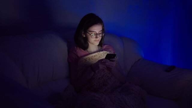 hapy girl sits on the sofa near the TV and uses the phone in the evening