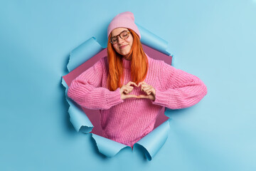 Romantic concept. Satisfied affectionate redhead woman does heart symbol or love sign shapes heart with fingers closes eyes with pleasure wears pink hat and sweater breaks through paper wall