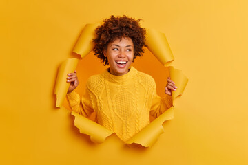 Glad ethnic girl with curly Afro hair smiles broadly concentrated aside giggles positively wears casual yellow sweater poses through ripped paper hole chuckles pleasantly notices something nice