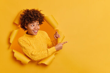 Try this! Cheerful pleasant looking young Afro American woman points away on copy blank space dressed in casual jumper breaks through paper wall gives recommendation asks to use good opportunity