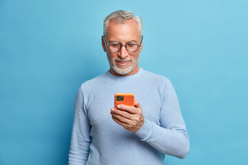 Photo of satisfied bearded man focused in smart phone surfs internet sends text messages in social networks uses modern technologies wears casual blue jumper poses indoor. Senior freelancer.