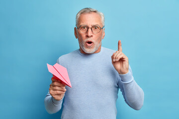 Surprised stunned bearded mature grey haired man points index finger up got excellent idea holds paper plane keeps mouth opened wears glasses and jumper poses against blue studio wall. Omg concept