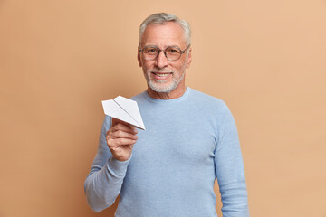 Satisfied confident mature grey haired man with smile holds handmade paper plane being sure in successful future wears glasses and blue jumper isolated over brown background. People and age concept