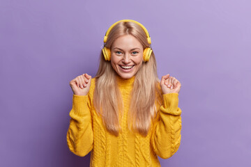 Pleased beautiful blonde teenage girl raises hands with happy expession smiles pleasantly wears headphones and listens music isolated over purple background. Listening pleasant melody or favorite song