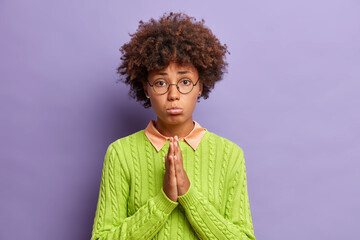Upset dark skinned woman with sad expression says sincerely please presses palms together in...