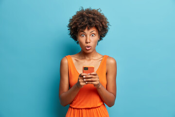 Studio shot of astounded startled curly haired woman wears dress holds modern cell phone looks with shocked expression uses free wifi reads message content isolated over blue studio background.