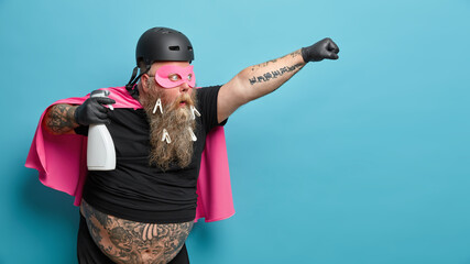Self confident brave man wears superhero costume raises arm holds cleaning detergent has big...