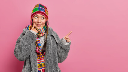 Pleased Asian woman with cheerful smile indicates away on copy space demonstrates blank empty place for your advertisement wears winter clothing isolated over pink bacground. Follow this direction