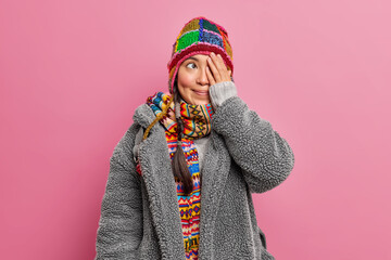 Playful positive young woman covers face with palm thinks about something wears winter clothing poses against pink studio wall. Lovely female model dressed in knitted hat scarf and fur grey coat