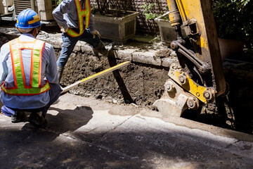 A backhoe and crew dig a utility trench for gas and other electrical utilities at a local community. Workers are measuring the width of the trench.