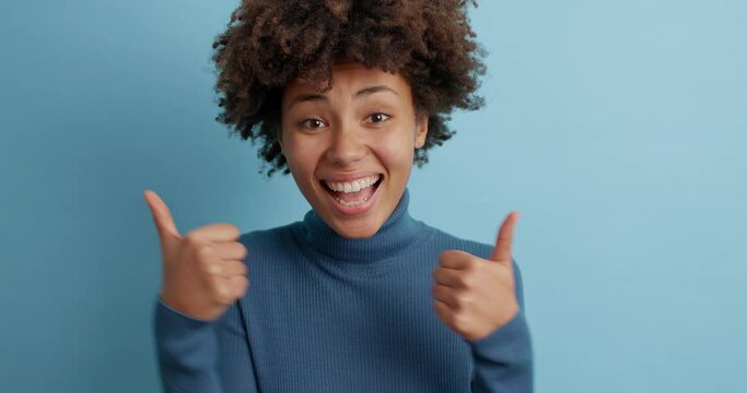 Motivated cheerful dark skinned woman makes thumb up gesture gives positive feedback approves you idea or likes something wears casual turtleneck poses against blue background. Body language