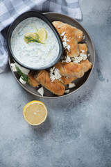 Filo mini rolls with feta cheese stuffing and tzatziki dip, above view on a light-blue stone background, vertical shot with copy space