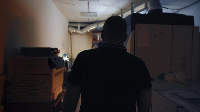 Workplace in office cubicle. Stock footage. Man walks across dark, small room to light-colored Desk in corner. Office worker works in copy room with document warehouse
