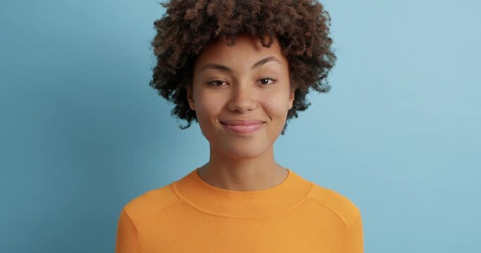 Satisfied young Afro American woman confirms something and nods head with acceptance gives positive reply smiles gently dressed casually poses against blue studio wall. Slow motion. Agreement concept