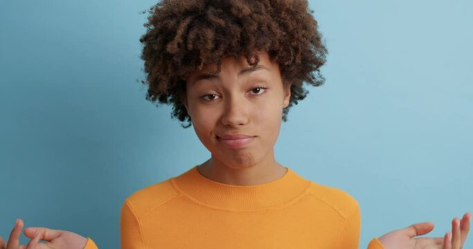 Indecisive brunette woman shrugs shoulders and raises palms in bewilderment cannot make choice stands doubtful indoor wears casual orange jumper being questioned isolated over blue background