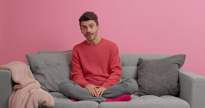 Serious guy reacts on something impressive looks with great wonder or surprisement at camera sits crossed legs on sofa watches scary movie poses in lotus pose against pink background at home