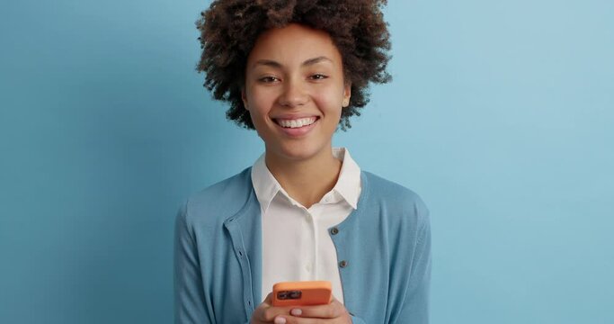 Curly haired young female smiles gladfully as sends text messages to friend holds modern cellular uses mobile phone surfs social networks wears white shirt and casual blue jumper poses indoor