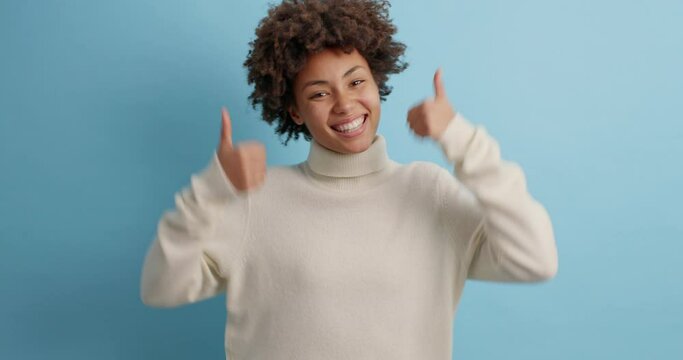 You can do it. Positive dark skinned woman raises thumbs up agrees with something or gives positive reply recommends advertisement likes good thing dressed in casual jumper poses indoor over blue wall