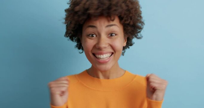 Cheerful optimistic Afro American woman makes winner gesture clenches fists with celebration and triumph smiles broadly dressed in casual orange jumper isolated over blue wall. Yeah dream come true