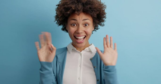 People emotions concept. Beautiful friendly looking Afro American woman waves palm in hello gesture says hi to you smiles broadly being pleased to see familiar face poses against blue background